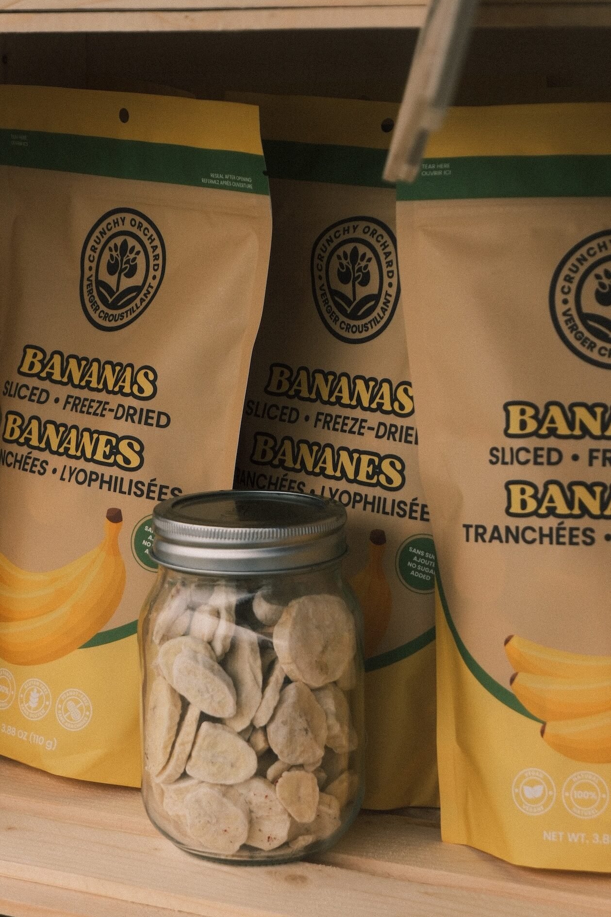 Bananes lyophilisées Verger Croustillant en sac et dans un pot en verre transparent montrant la texture, sur un présentoir en bois au marché fermier